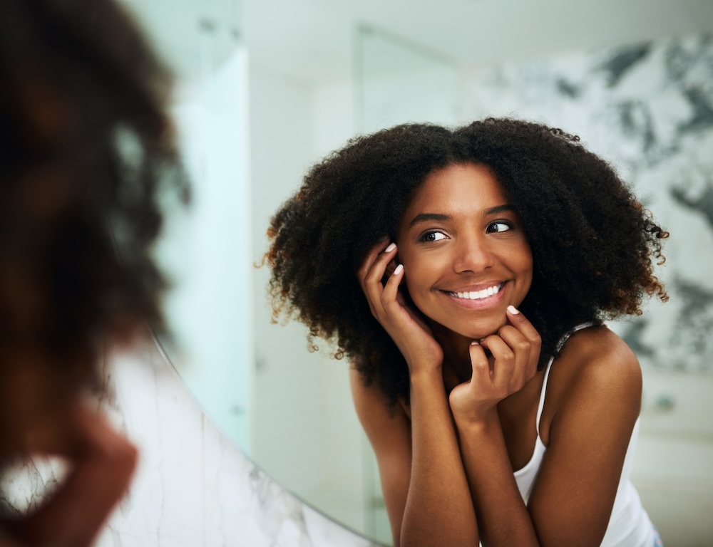 A young woman of color looks in the mirror, smiling after her professional teeth cleaning in Waxhaw.