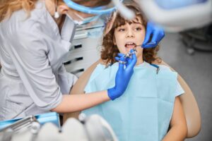 A child patient lies on a treatment bed as a female Dogwood Dental Spa provider, wearing blue gloves, leans over him and cleans his teeth. She may recommend dental sealant in Waxhaw to protect his teeth from cavities.